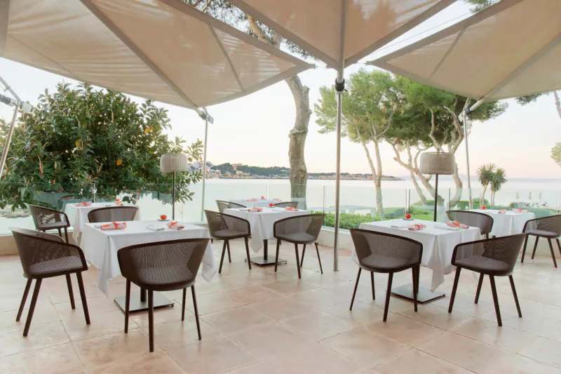 Outdoor terrace dining at ARRELS by Marga Coll with wicker chairs, white tablecloths, under beige umbrellas, sea view at sunset.