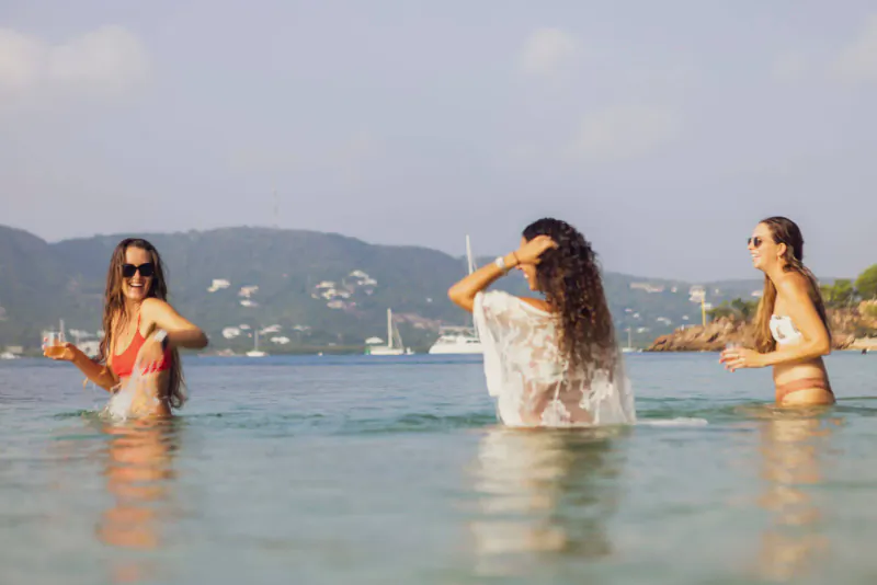Three women in bikinis and cover-ups splashing in turquoise sea at sunny Antigua beach with hills and sailboats.
