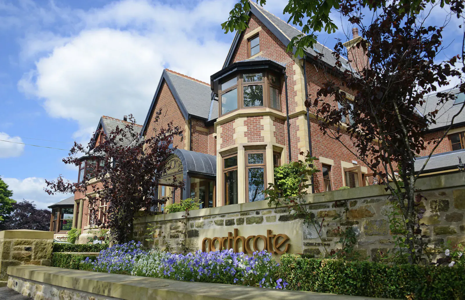 Northcote Hotel, a grand Victorian brick mansion with bay windows, surrounded by trees, flowers, and stone wall sign.