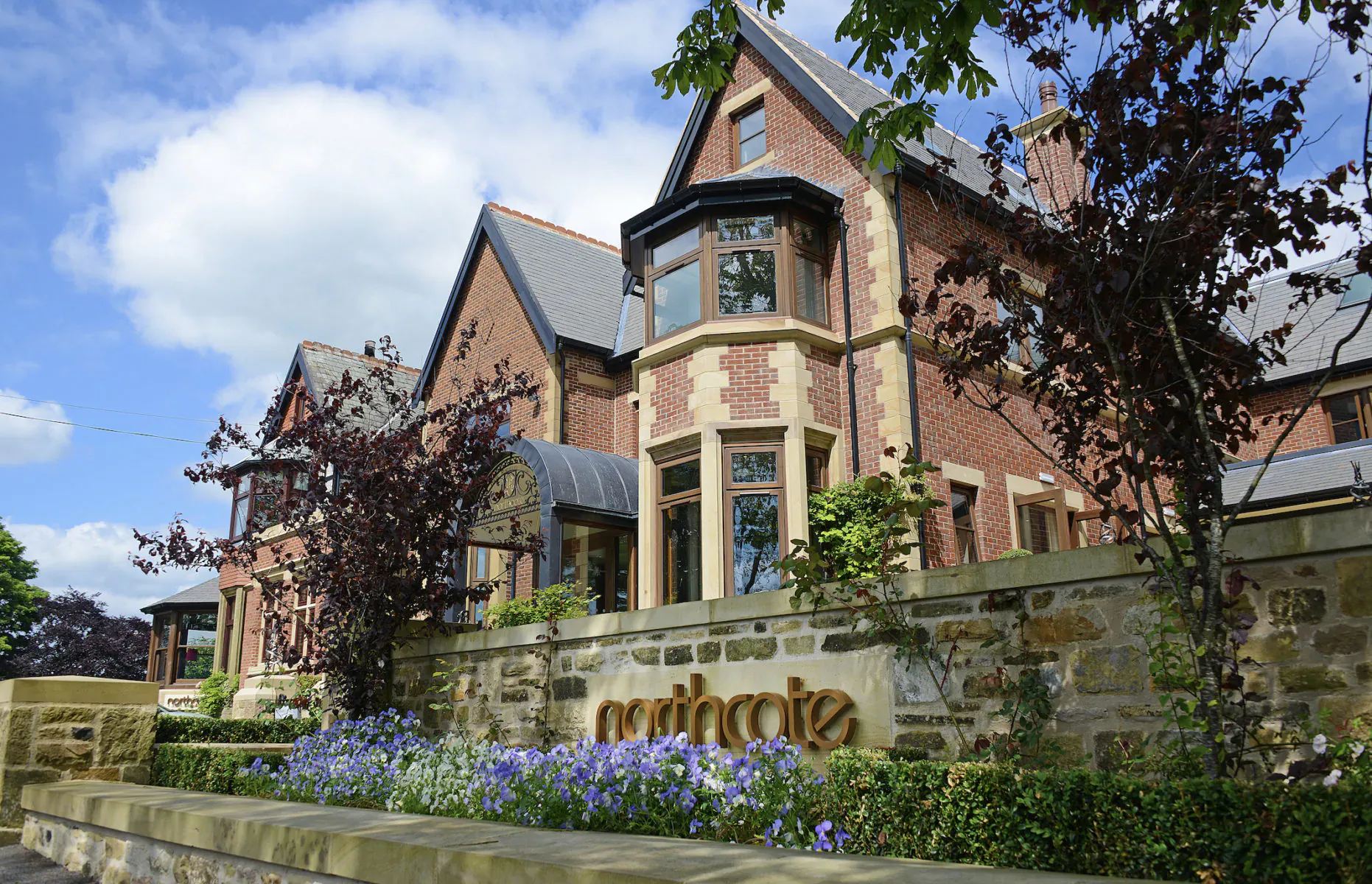 Northcote Hotel, a grand Victorian brick mansion with bay windows, surrounded by trees, flowers, and stone wall sign.
