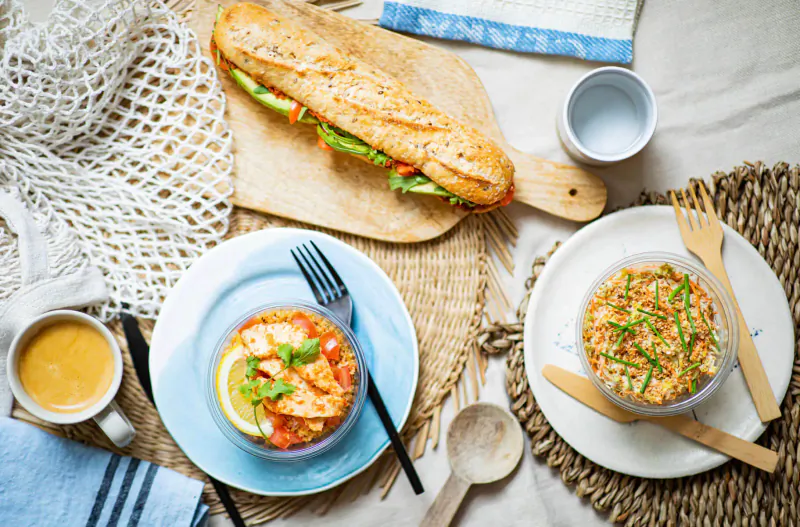 Picnic-style spread: baguette with veggies on board, turmeric latte, tomato feta salad, and grain bowl with utensils.