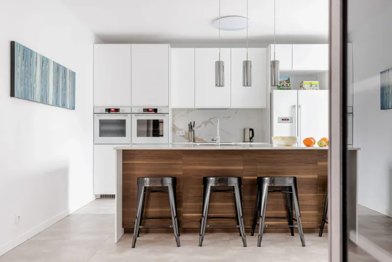 Modern white kitchen with wooden island, black stools, pendant lights, oranges in bowl, and blue abstract wall art.