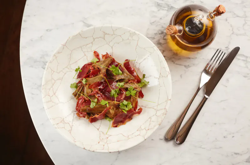 Plate of thinly sliced beef with herbs in white bowl, beside oil bottle and cutlery on marble table