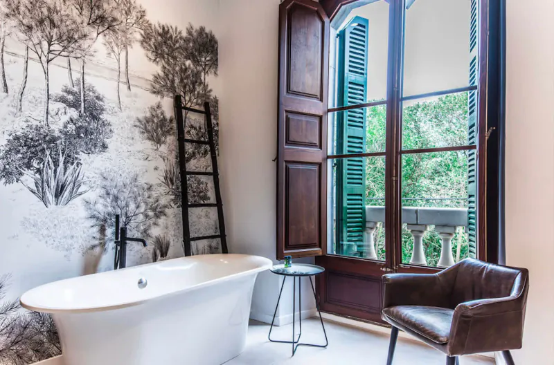 Elegant bathroom in L’Avenida Sóller Hotel with freestanding tub, ladder, leather chair, tree mural, and arched green shutters opening to garden.