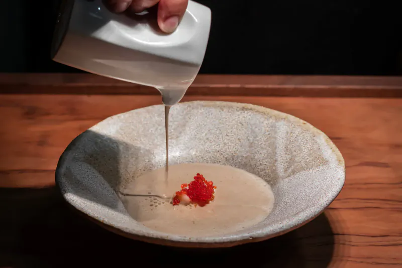 Hand pouring white sauce from pitcher into bowl of creamy soup with red garnish on wooden table