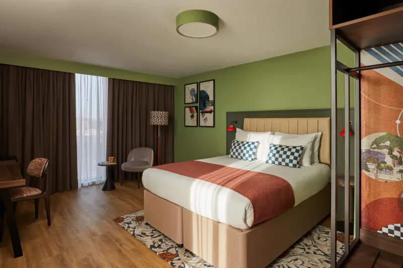 Cozy green bedroom in Hotel Indigo Coventry with king bed, checkered pillows, art, lamp, and window view.