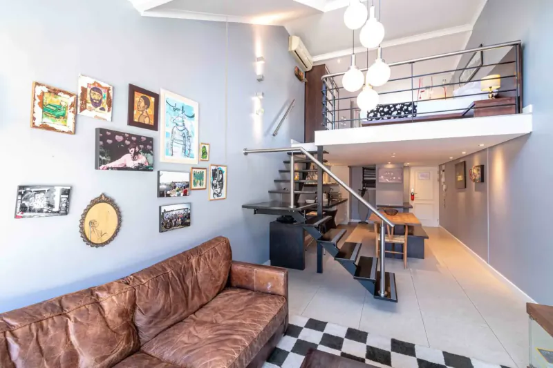 Modern loft room at Hippo Boutique Hotel with leather sofa, black-and-white tile floor, spiral stairs to mezzanine bed, 'PIZZA' sign, eclectic wall art.