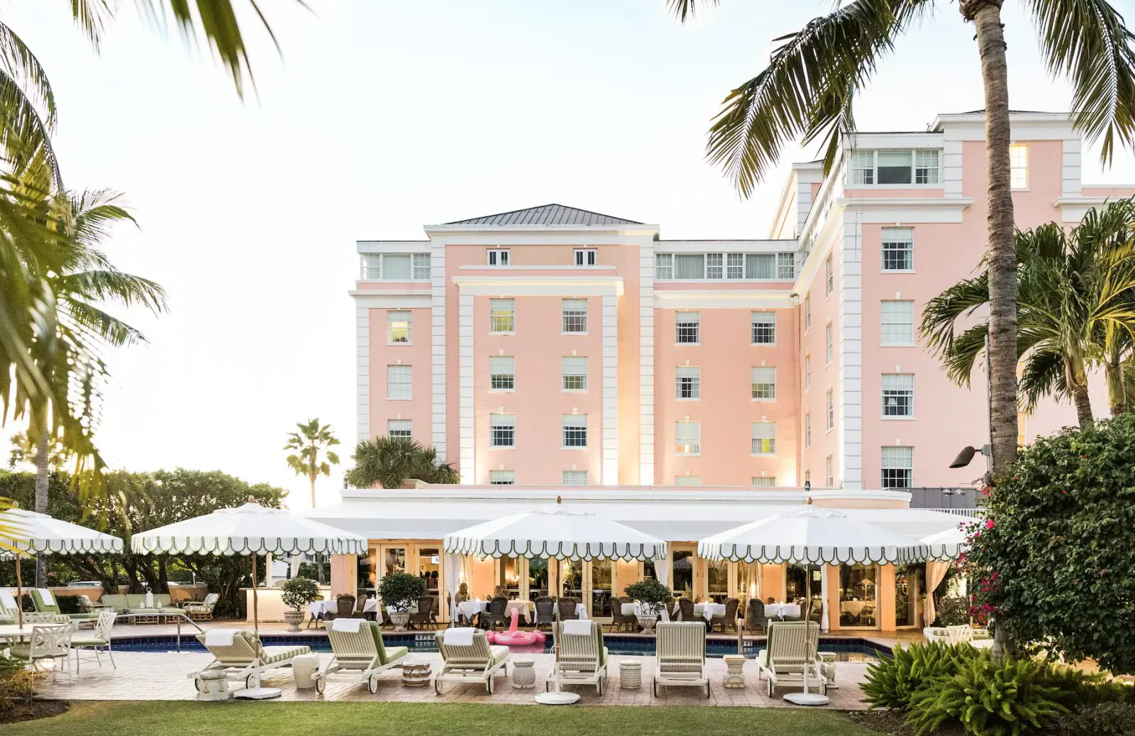 Pink Palm Beach hotel with pool, lounge chairs under striped umbrellas, surrounded by palm trees and gardens.