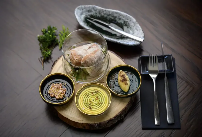 Elegant plated dish with poached fish in glass bowl, garnished with herbs, small sauces, butter, chopsticks, and forks on wooden board