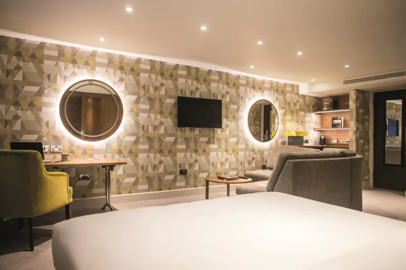 Modern hotel room with geometric wallpaper, round mirrors, TV, yellow chair, desk, sofa, and bed at Bristol Aztec Hotel.