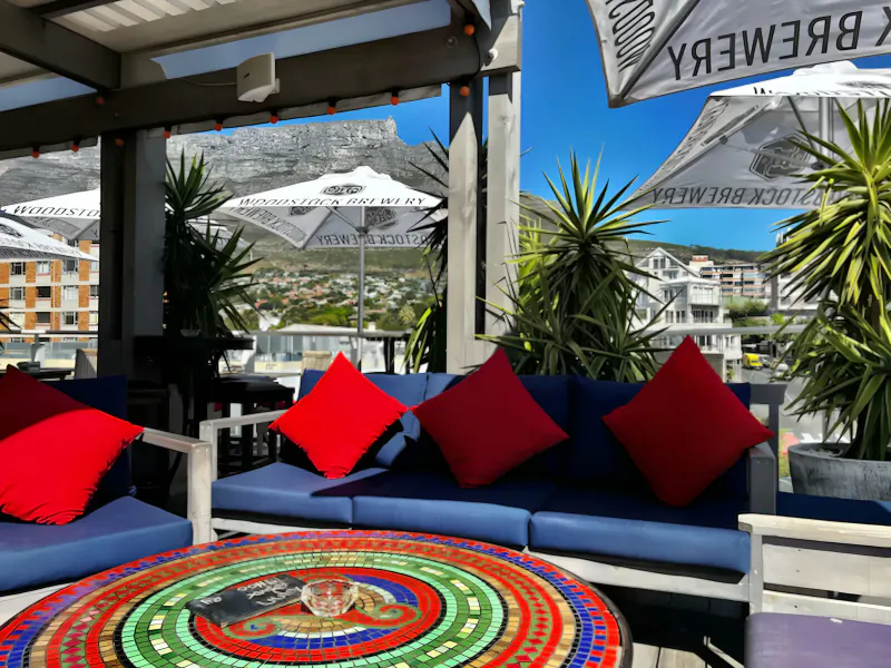 Outdoor Kloof Street lounge in Cape Town with blue sofa, red cushions, mosaic table, brewery umbrellas, Table Mountain view.