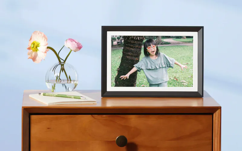 Aura smart photo frame on wooden nightstand with notepad, pink flowers in vase; shows joyful girl running by tree.