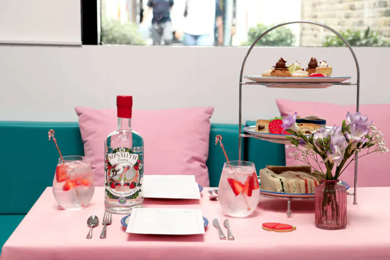 Pink high tea table with gin cocktails, tiered stand of cakes and sandwiches, flowers, by window.