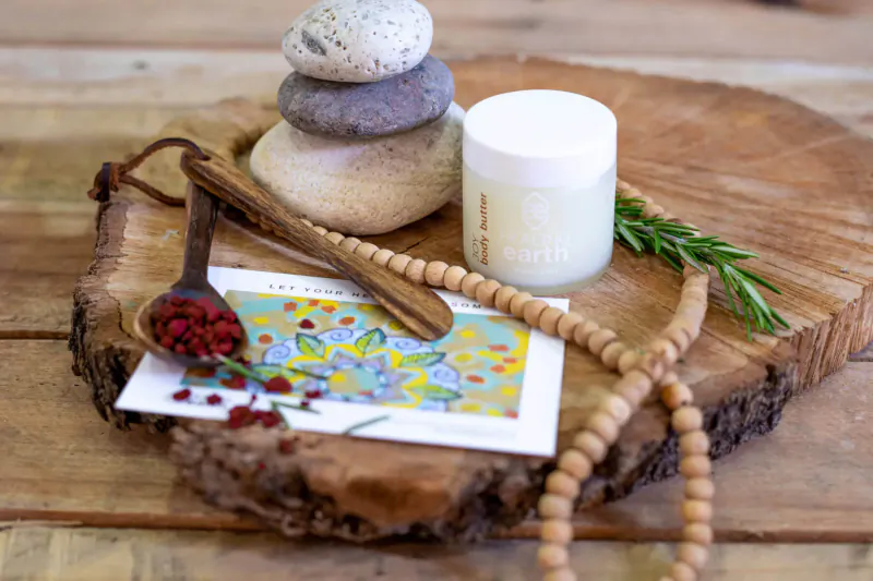 Wellness still life on wood slice: stacked stones, white cream jar, wooden spoon, beaded necklace, rosemary, floral card with berries.