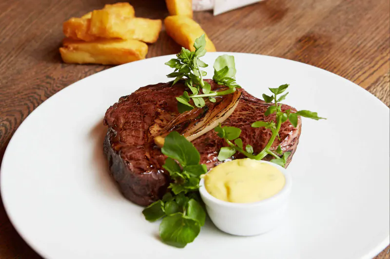 Medium-rare steak garnished with herbs and onions on white plate with béarnaise sauce and fries beside, on wooden table.