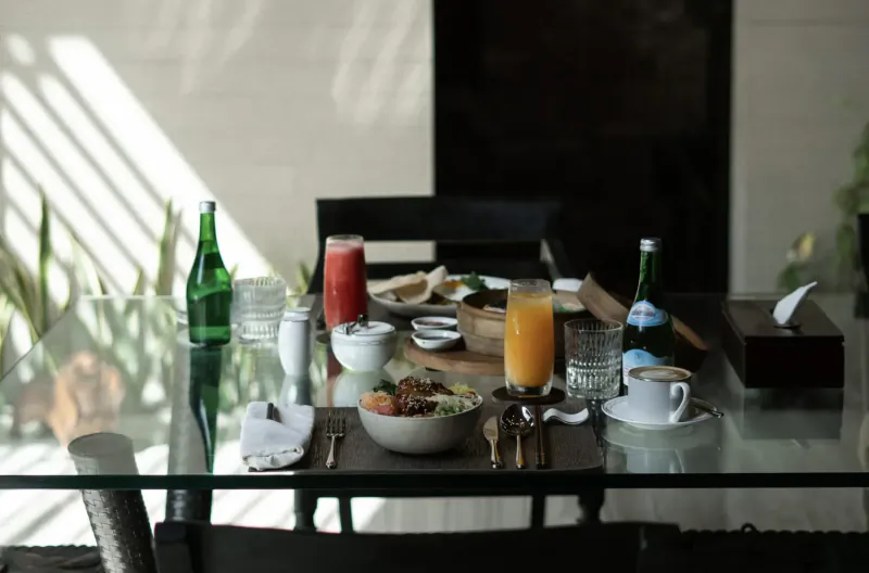Glass dining table set with fresh salad bowl, orange juice, green bottles, coffee cup in sunlit villa room