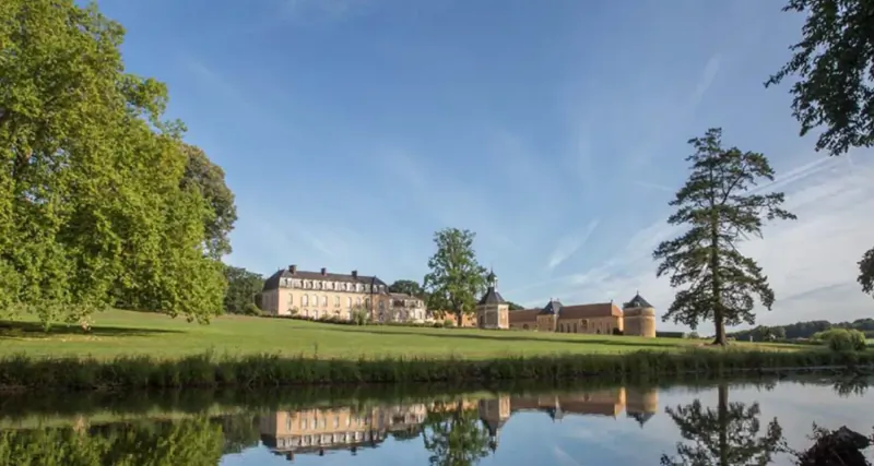 Grand château with manicured gardens, trees, and serene pond reflection under blue sky