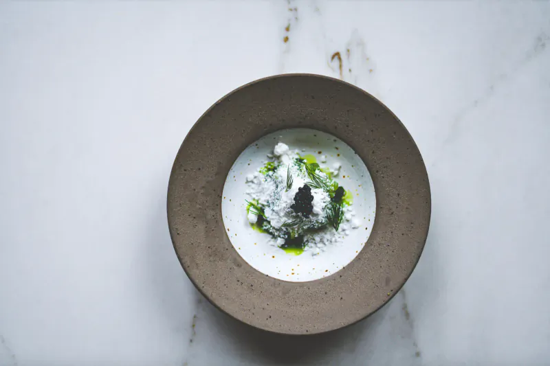 Creamy white soup with green herbs and powder in brown bowl on marble surface