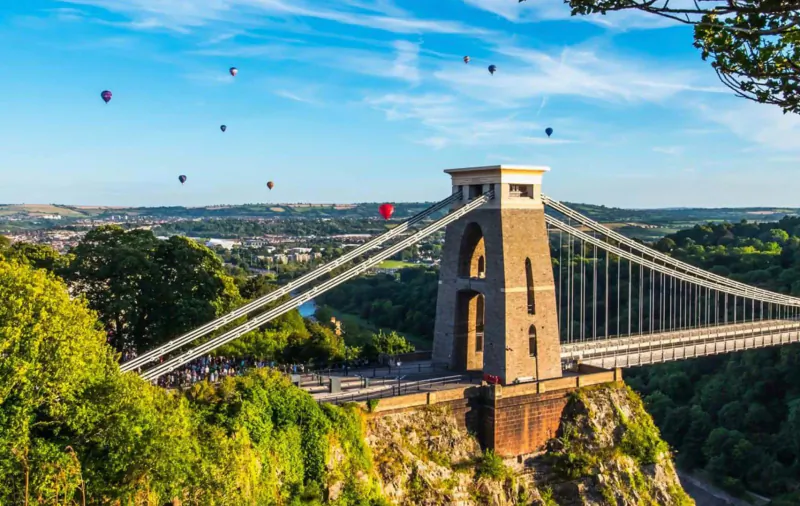 Clifton Suspension Bridge in Bristol with colorful hot air balloons floating above, green hills and blue sky.