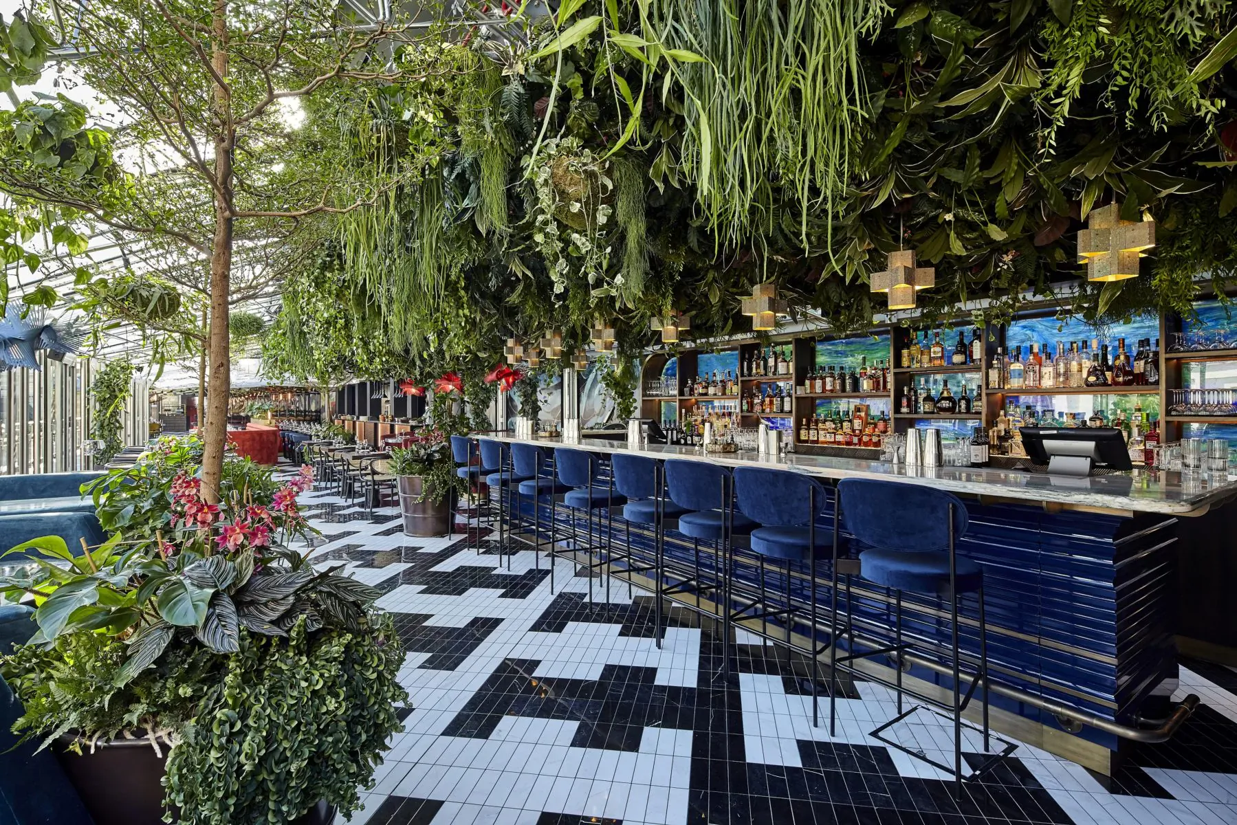 Sushi Samba lush greenhouse bar with hanging plants, blue stools, checkered floor, and stocked shelves