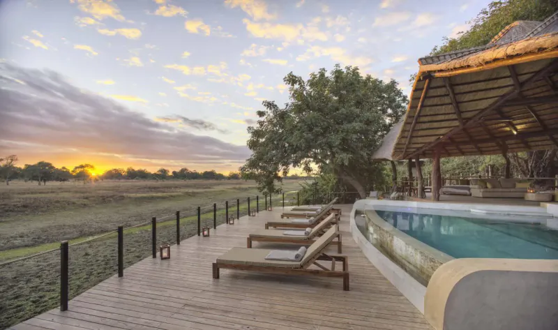 Lion Camp's infinity pool and lounge chairs on wooden deck at sunset, overlooking African savanna river.
