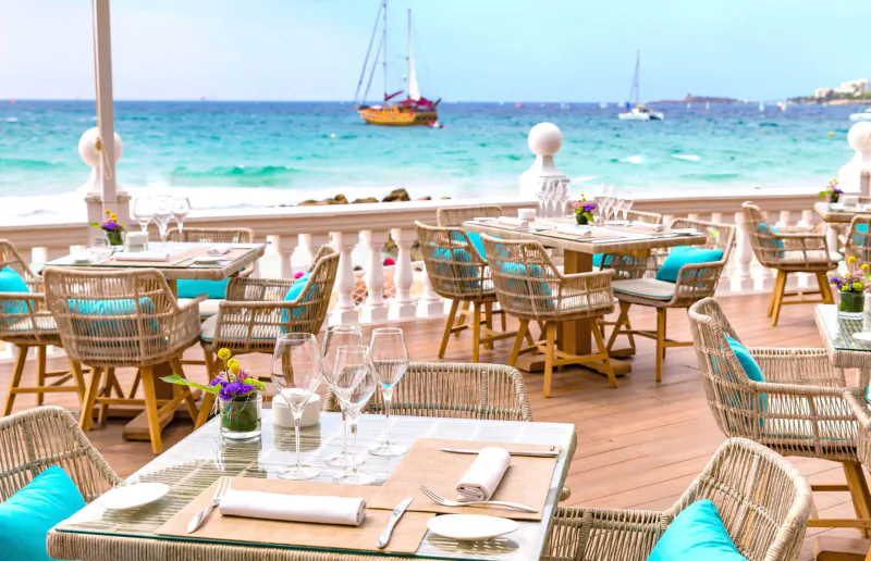 Luxury beachfront restaurant at Nixe Palace with turquoise wicker chairs, set tables, sailboats on sea beyond.