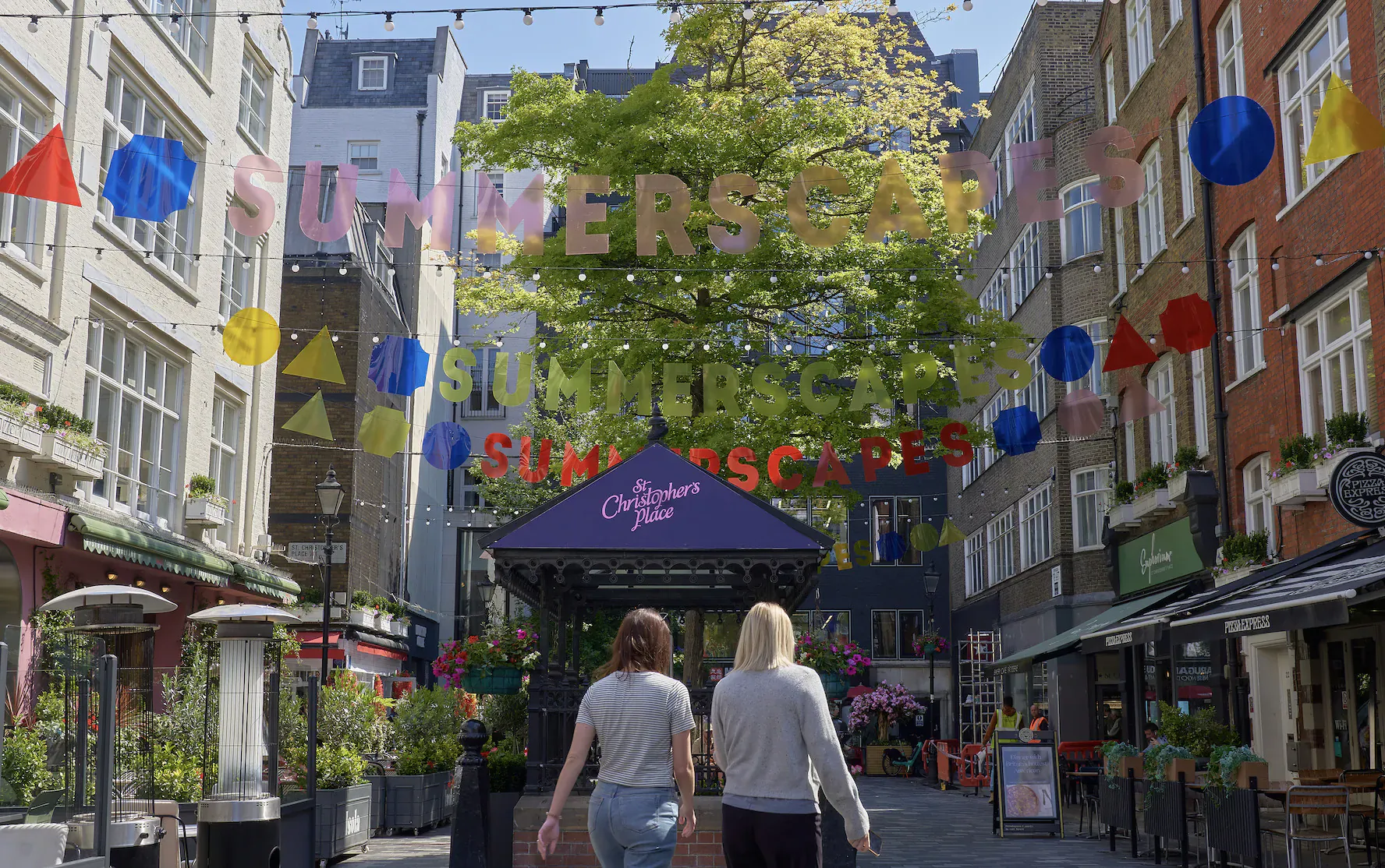 Two women walking past 'Summer Escapes' arch and purple pavilion in London's colorful St. Christopher's Place street.