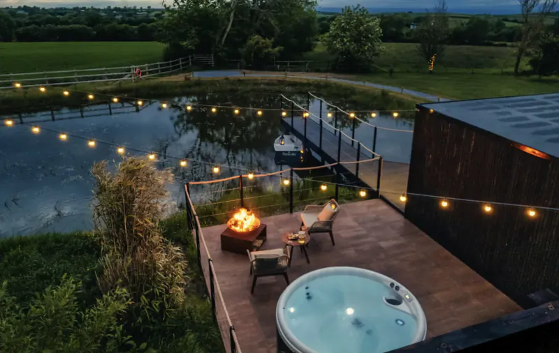 Aerial view of The Boathouse luxury lodge deck with hot tub, fire pit, chairs by pond at dusk in Welsh countryside.