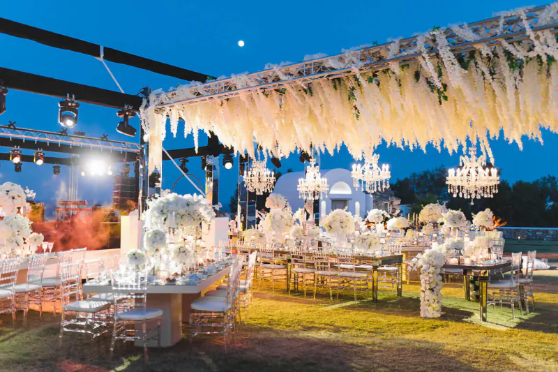 Elegant outdoor wedding reception in Greece at dusk with wisteria-draped pergola, chandeliers, and white floral dining tables on lawn