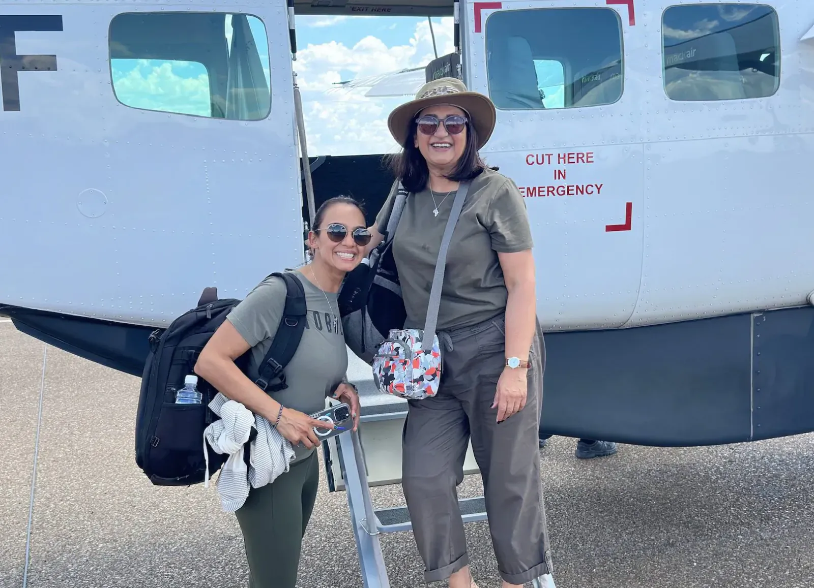 Two smiling women in hats and backpacks stand on airstair by airplane with 'CUT HERE IN EMERGENCY' sign, sunny day.