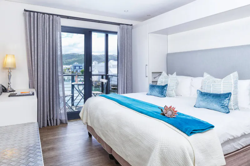 Luxurious hotel bedroom with king bed, blue runner and protea flower, balcony overlooking harbor and mountains.