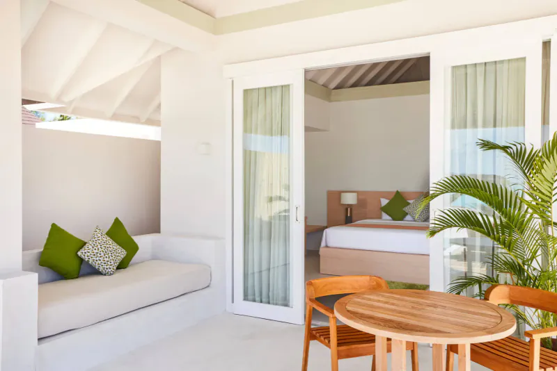 Bright tropical villa interior with open doors to bedroom, green pillows on white couch, potted palm, wooden table and chairs.