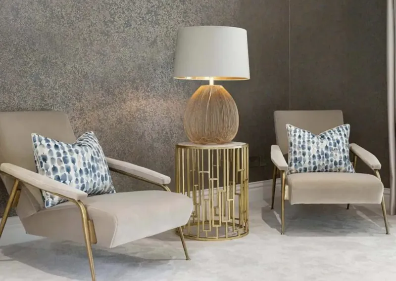 Two cream armchairs with blue pillows flank a tall rattan-shade lamp on gold geometric table in elegant gray room.