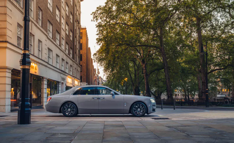 Silver Rolls-Royce sedan parked on London street at dusk amid trees and historic buildings.