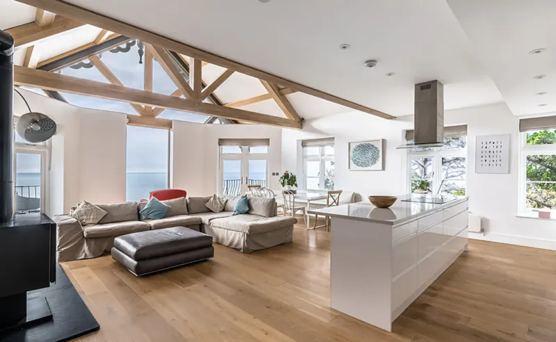 Spacious open-plan living room and kitchen in Tors Park Devon, with oak beams, sea-view windows, white island, beige sofas, and wood stove.