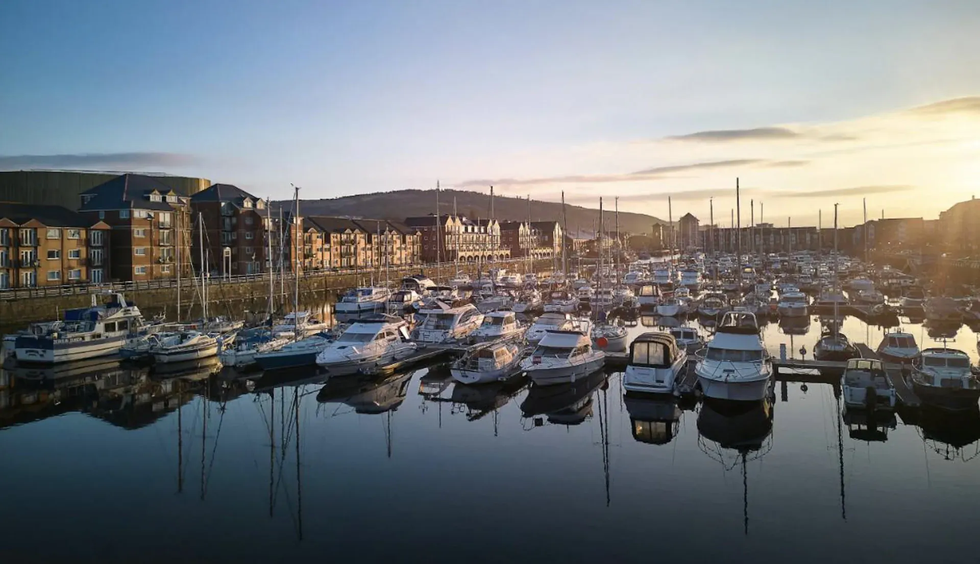 Aerial view of marina with docked yachts reflecting in calm water at sunset, modern apartments and hills in background near Delta Hotel Swansea.