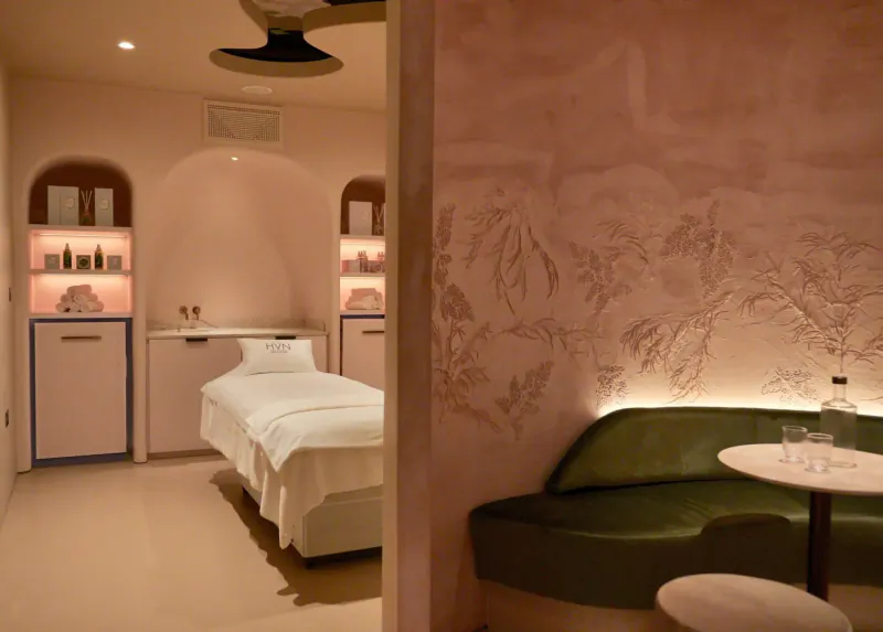 Elegant HVN spa room in Knightsbridge with white massage bed, green curved sofa, arched shelves with products, and leafy pink walls.