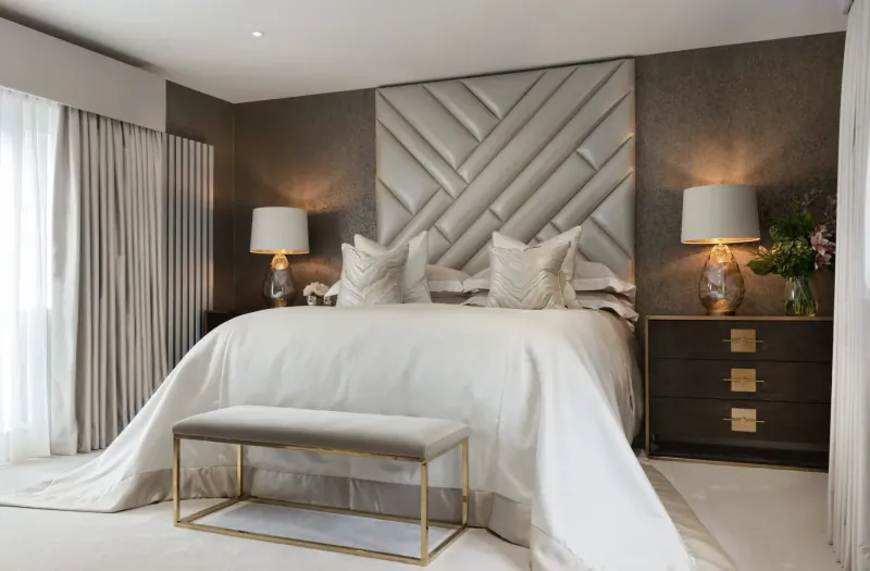 Elegant bedroom with white tufted headboard, king bed with white linens, bedside lamps, flowers, bench, and drapes.