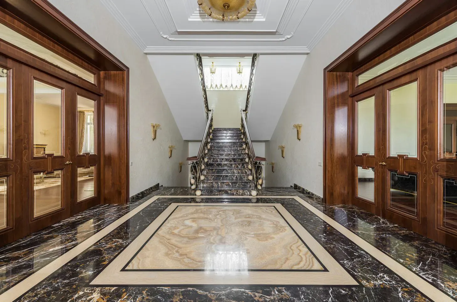 Luxurious grand entrance hall with ornate marble floor and sweeping wooden staircase framed by double doors.