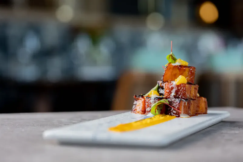Stacked glazed pork belly cubes with mango and microgreens on yellow sauce, Southeast Asian cuisine