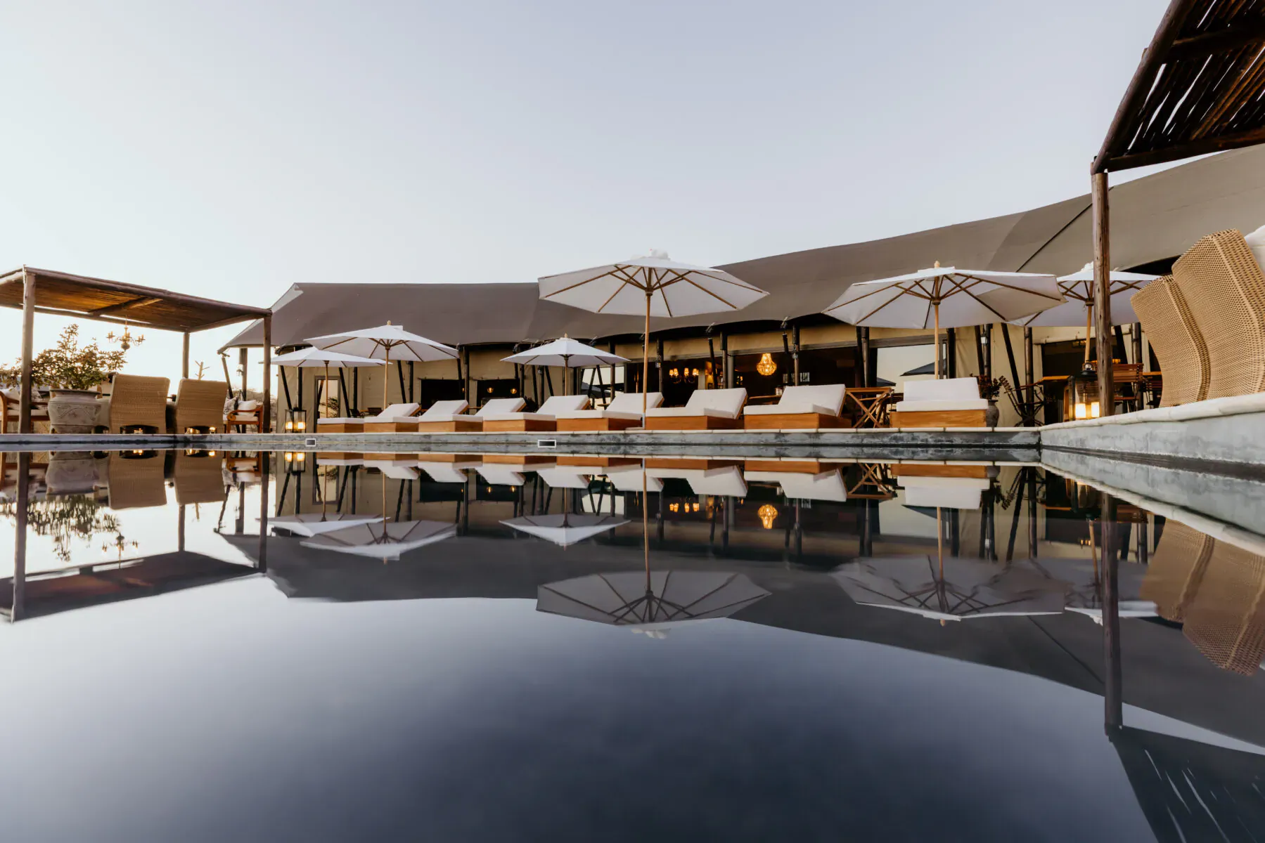 Infinity pool at Thornbush Tented Camp reflecting luxury loungers, umbrellas, and thatched safari tents at dusk.