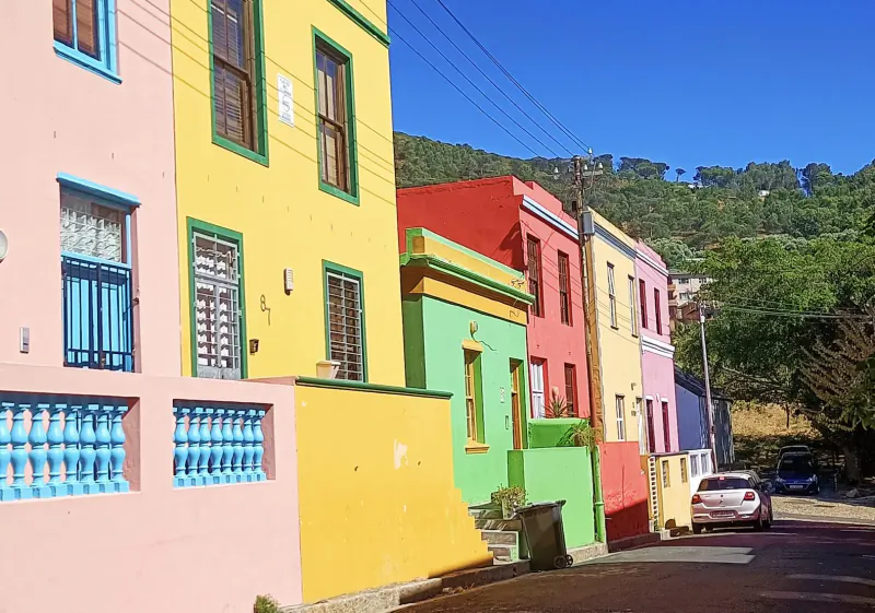 Colorful Bo-Kaap houses in Cape Malay District, vibrant pink, yellow, green, blue facades lining steep street with car, against green hillside and blue sky.