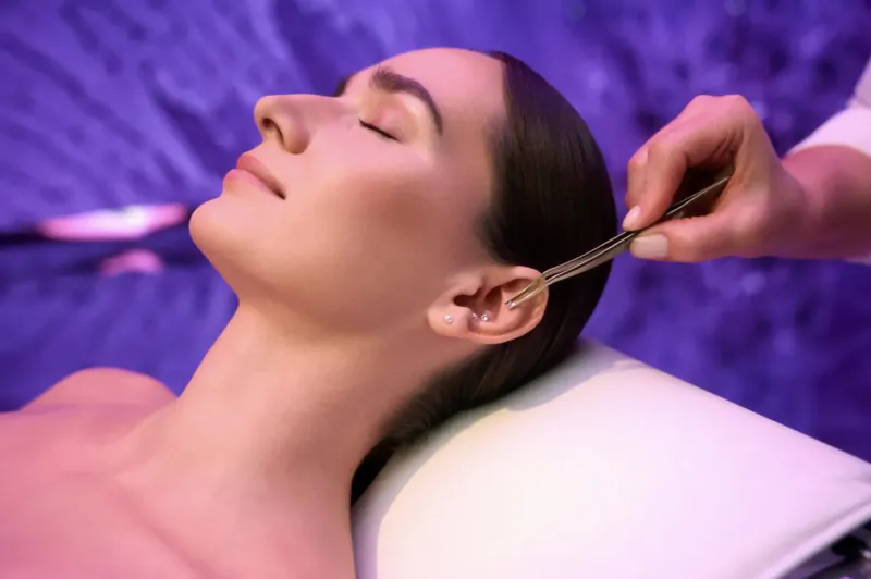 Woman lying relaxed on spa bed as therapist uses ear candles in purple-lit room