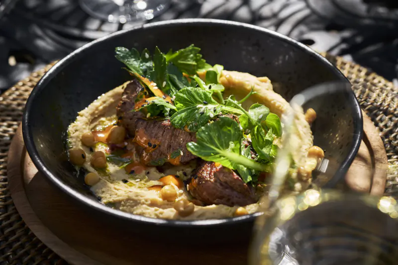 Black bowl of hummus topped with seared beef slices, chickpeas, greens, poured with white wine on outdoor table.