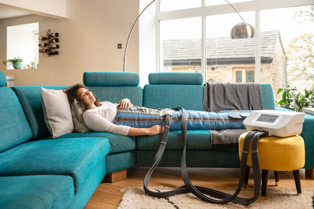Woman lying on teal sofa using Body Ballancer massage machine with leg wraps and control unit connected by tubes