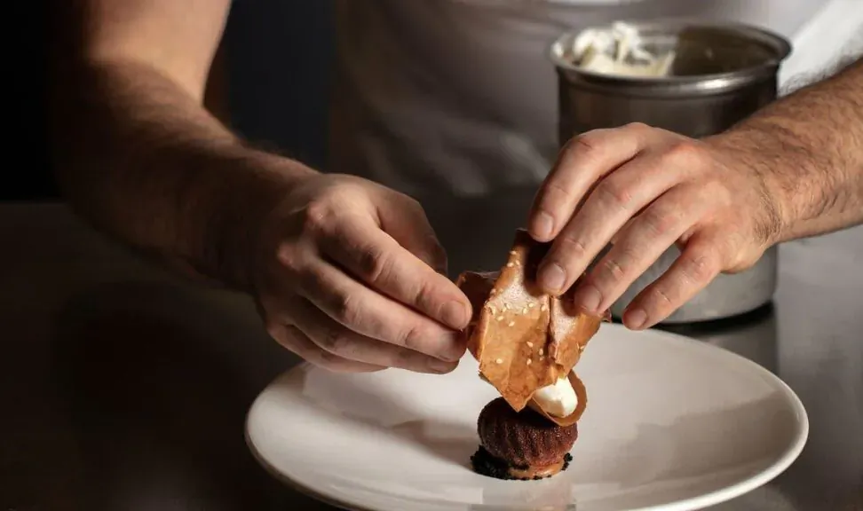 Chef's hands placing delicate wafer atop chocolate mousse with cream on white plate