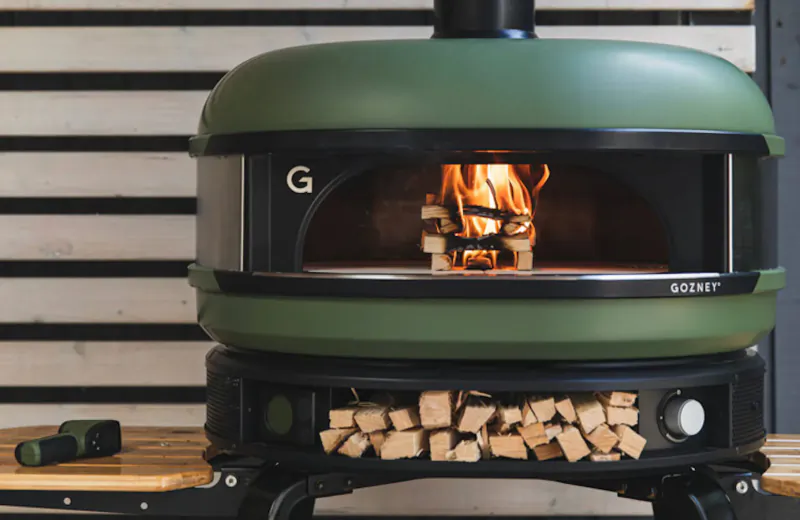Green Gozney wood-fired pizza oven with flames inside, firewood stack, on wooden base against white wall.
