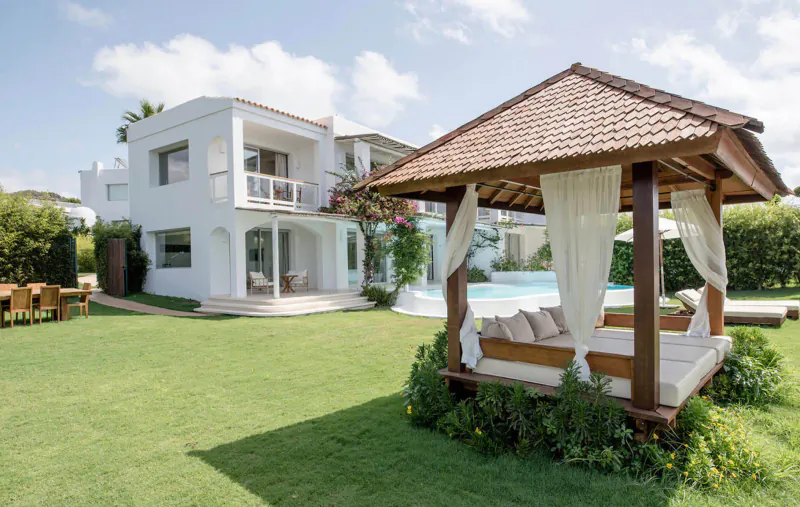White modern Ibiza villa with pool, wooden gazebo daybed with cushions, lush green lawn and gardens.