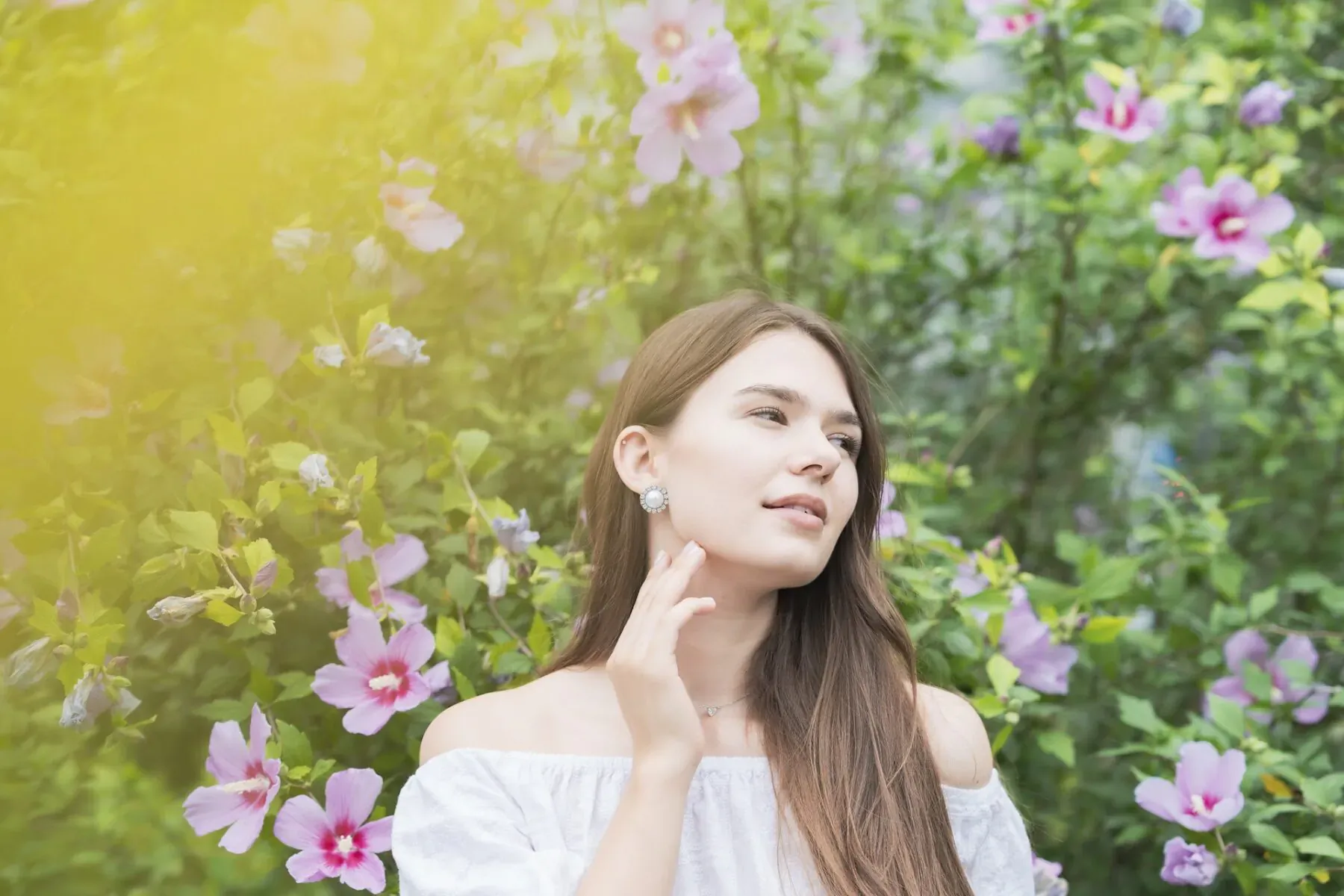 Young woman with long brown hair touching her smiling face, wearing off-shoulder white top amid pink hibiscus flowers