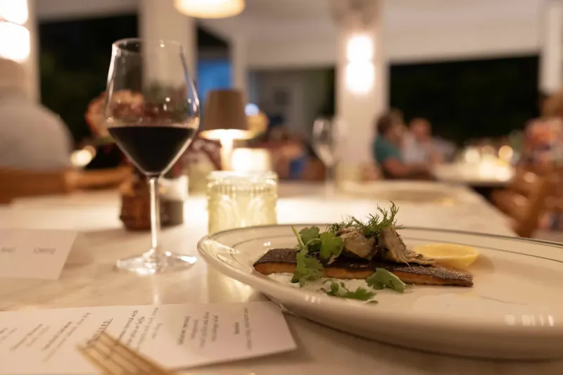 Close-up of red wine glass beside white plate of seared scallops with herbs and lemon on restaurant table.